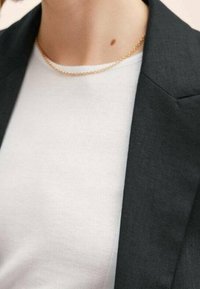 Černý límec blejzru vedle bílé, texturované halenky; jemný zlatý řetízek viditelný na krku. Mezi subtilními detaily se nachází malá znaménka.