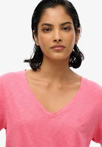 Camiseta rosa de cuello en V fabricada en un tejido ligero con una sutil textura jaspeada. Presenta un pequeño logo en el pecho. Manga corta y corte holgado.