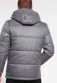 Mand iført en grå polstret vinterjakke med hætte, set bagfra mod en ensfarvet lys baggrund.