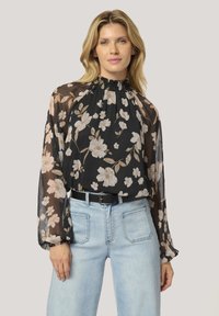 Blusa nera floreale con maniche lunghe trasparenti e scollatura arricciata. Presenta grandi fiori color crema ed è abbinata a jeans azzurri chiari.