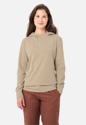 Frau mit langem braunem Haar, die ein beiges Kapuzenoberteil mit langen Ärmeln und rostfarbene Hosen trägt, steht mit einer Hand in der Tasche.