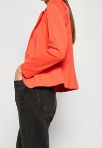 Blazer corail en tissu léger, avec une fermeture à un seul bouton, une coupe ajustée et un ourlet incurvé. Associé à un jean foncé.