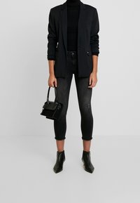 Blazer negro sobre un cuello de tortuga negro, combinado con jeans de mezclilla oscuros con dobladillos deshilachados. Sosteniendo un pequeño bolso negro. Botines negros.