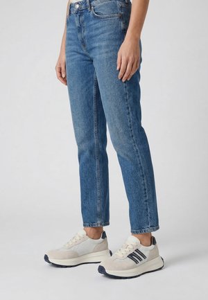 Underkrop iført straight-fit blå jeans og hvide sneakers med sorte striber, stående på en ensfarvet lys baggrund.