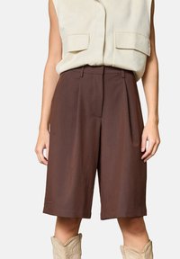 Brune skræddersyede shorts lavet af et letvægtsstof, med et plisseret frontdesign og en midt-lang længde. Båret med en beige ærmeløs top.