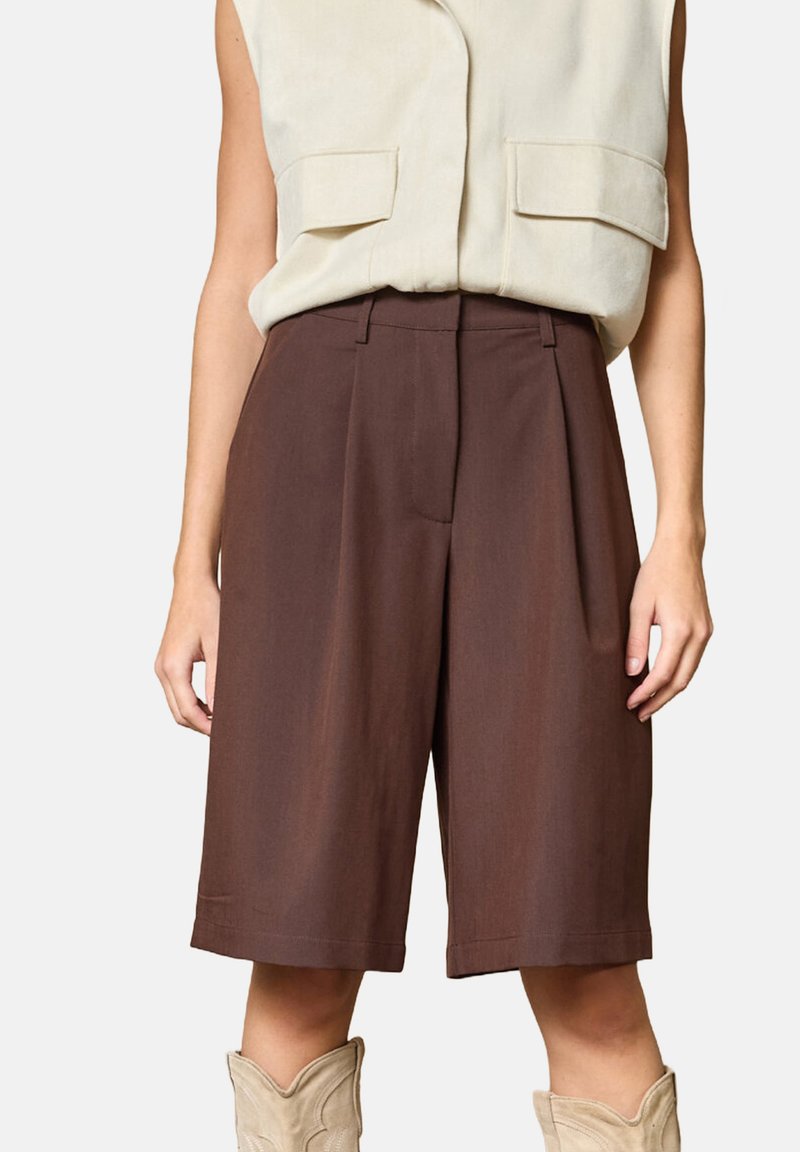 Brune skræddersyede shorts lavet af et letvægtsstof, med et plisseret frontdesign og en midt-lang længde. Båret med en beige ærmeløs top.