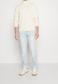 Man som bär en krämfärgad polotröja, ljusblå jeans med uppvikt ben och vita sneakers som står mot en slät vit bakgrund.