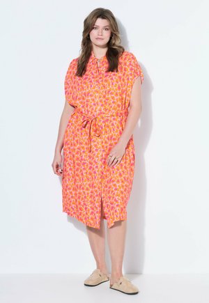 Kvinde med løst brunt hår iført en orange og pink mønstret knælængde bælte kjole og beige slip-on sko, stående mod en hvid baggrund.