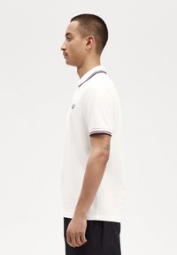 Witte polo met een geribbelde kraag met rode en marineblauwe strepen. Klein logo op de borst. Model weergegeven in profiel.