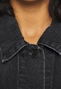 Närbild av en persons nacke och övre bröst som har på sig en mörk denimjacka med en stor krage och en enda svart knapp knäppt.