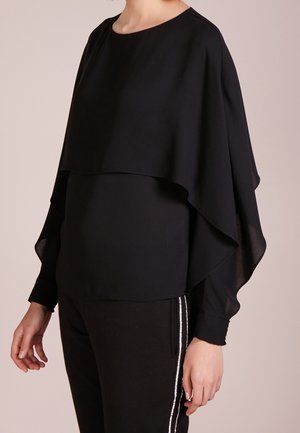Haut noir superposé avec un design fluide de type cape, fabriqué en tissu léger. Présente des manches longues et un col rond.