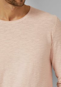 Man met lichtroze, lang mouw, getextureerd gebreid shirt met een ronde hals tegen een effen grijze achtergrond.