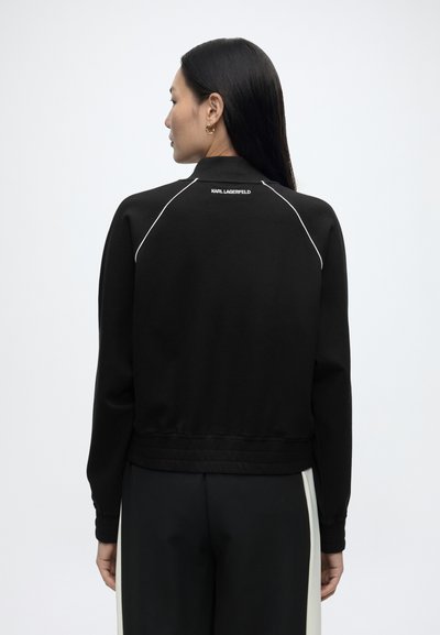 Femme aux longs cheveux noirs portant une veste noire avec des liserés blancs et le texte « Karl Lagerfeld », vue de dos sur un fond uni.