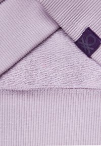 Un tessuto lilla presenta una texture a coste nella parte superiore e un design a luppolo sotto, con un piccolo etichetta viola che mostra un logo.