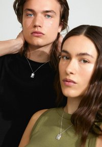 Collar con colgante de plata usado sobre una camisa negra, combinado con una blusa sin mangas verde. Ambas figuras están en primer plano, con fondos neutros.