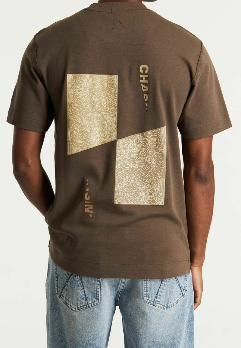 Personne portant un t-shirt marron avec deux impressions de cartes topographiques beiges et un texte partiel dans le dos, associé à un jean bleu clair.