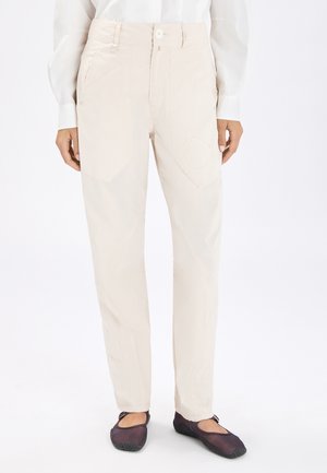 Camicia bianca a maniche lunghe infilata in pantaloni color crema a vita alta con tasche applicate, abbinata a scarpe slip-on nere, su sfondo bianco.