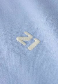 Ljust blått textil med en fint vävd textur och en krämfärgad broderad "21"-logotyp i modern stil.
