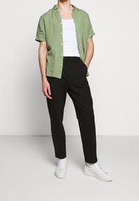 Vihreä pellavapaita, jossa on lyhyet hihat ja valkoinen toppi alla, yhdistetty mustiin housuihin ja valkoisiin lenkkareihin. Siistit linjat ja rento sovitus.