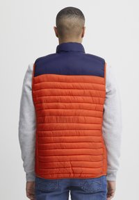 Polstret vest med orange nederste sektion og mørkeblå øverste sektion. Har en høj krave og quiltet tekstur. Båret over et lysegråt tøj.