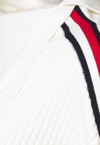 Vêtement tricoté blanc avec texture côtelée, présentant des accents de bandes noires et rouges le long de la couture de l'épaule. Détail de design notable : une couture contrastée.