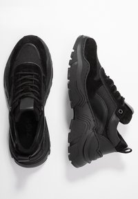 Baskets noires présentant un mélange de textures lisses et en suède, semelle en caoutchouc épaisse, bout arrondi et fermeture à lacets. Détails comprenant un onglet de tirage.