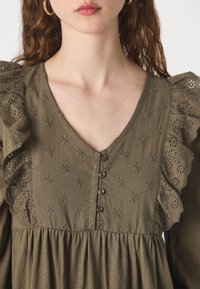 Ελιόχρωμο φόρεμα με V λαιμόκοψη, διακοσμητικά κουμπιά, κεντημένα λουλουδάτα σχέδια και ραμμένα δαντελένια περιθώρια στους ώμους.