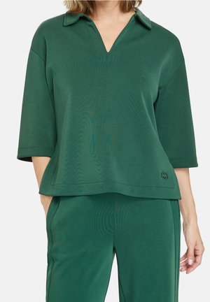 Kvinde iført løs mørkegrøn bluse med krave og matchende bukser, hænder afslappet ved siderne, beskåret udsigt fra nakke til lår.