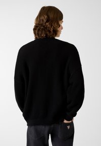 Schwarzer Strickpullover mit Wabenstruktur, überschnittenen Schultern und gerippten Bündchen. Kombiniert mit dunkler Jeans, Ansicht von hinten.