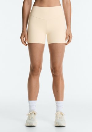 Frau trägt beige hoch taillierte, eng anliegende Shorts, weiße Kniestrümpfe und beige Sneakers, steht vor einem schlichten hellen Hintergrund.
