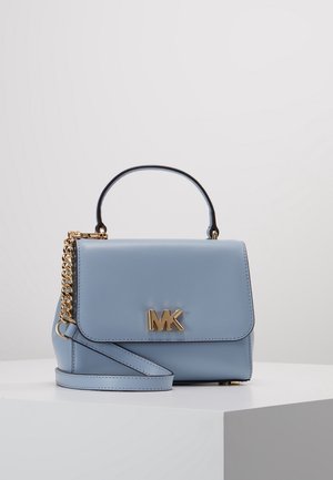 Lichtblauwe leren handtas met gouden "MK"-logo, bovenhandvat en afneembare schouderriem, rustend op een wit voetstuk tegen een effen achtergrond.