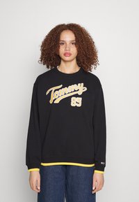 Svart sweatshirt med gul "Tommy" och "85" tryck, ribbade ärmar med gula accenter och mjuk tygtextur.