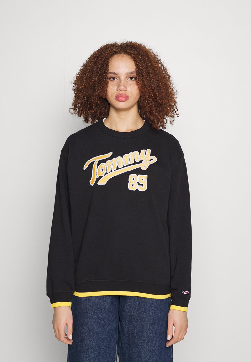 Svart sweatshirt med gul "Tommy" och "85" tryck, ribbade ärmar med gula accenter och mjuk tygtextur.