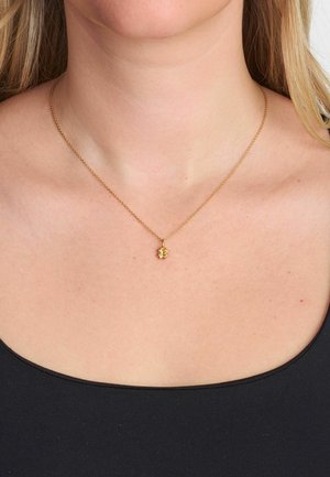 Złoty naszyjnik z małym wisiorkiem w kształcie kwiatka, z delikatnymi detalami. Noszony na czarnej bluzce, podkreślając wzór naszyjnika.