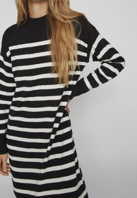 Randig svart och vit tröjdress, stickad kvalitet, hög hals, långa ärmar, med en avslappnad passform och slitsar på sidorna för ökad rörelsefrihet.