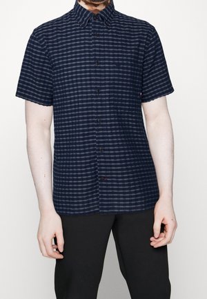Man draagt een kort mouw marineblauw en zwart geruit overhemd met knopen en zwarte broek, armen ontspannen langs het lichaam.