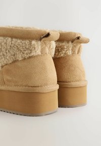 Beige ruskind ankelstøvler med en blød, uldforet krave og en kraftig, gummibelagt platformssål. Har en syet detaljering langs kanten.