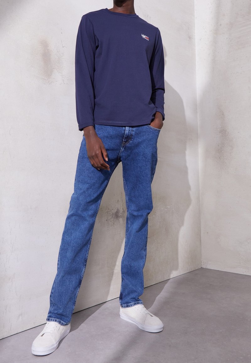 Marinblå långärmad skjorta med liten logotyp, kombinerad med blå denimjeans och vita sneakers. Slät tygkvalitet och avslappnad passform, står mot en ljus vägg.