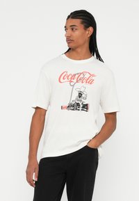 Valge puuvillane T-särk, millel on punane Coca-Cola logo ja graafika cowboy-kangelasest, kes istub limonaadipudeli peal. Lühikesed varrukad, mugav lõige.