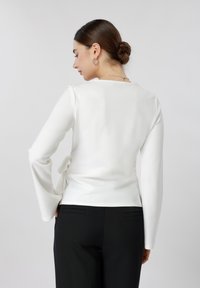 Γυναίκα με σκούρα μαλλιά σε κότσο, που φορά ασημένια σκουλαρίκια κρίκους, λευκό μακρυμάνικο μπλουζάκι με φιόγκο στο πλάι και μαύρο παντελόνι, στραμμένη προς τα πίσω σε απλό φόντο.