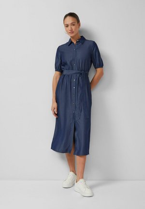 Kobieta ubrana w granatową sukienkę midi na guziki z paskiem w talii i bufiastymi rękawami, w białych tenisówkach, stojąca na tle gładkiej ściany.