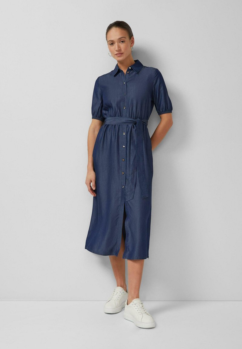 Kobieta ubrana w granatową sukienkę midi na guziki z paskiem w talii i bufiastymi rękawami, w białych tenisówkach, stojąca na tle gładkiej ściany.