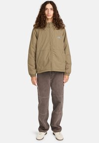 Jongere met lang krullend haar die een beige ritsjack, losse bruine broek en witte sneakers draagt, staand tegen een witte achtergrond.