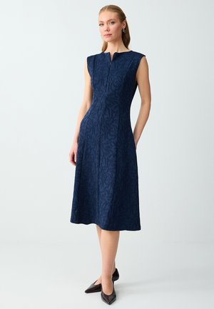 Femme debout, les mains dans le dos, portant une robe midi sans manches bleu marine avec un motif subtil de feuilles et des chaussures noires à bout pointu.