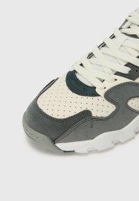 Sport shoes with a grey and cream upper, featuring perforated detailing, textured materials, and white laces against a light background.