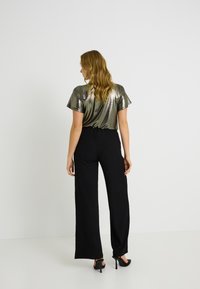 Un haut à manches courtes vert métallique associé à un pantalon large noir. Le tissu a un subtil éclat, créant un contraste texturé. Des escarpins noirs complètent le look.