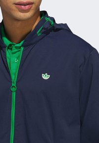 Chaqueta con capucha de color azul marino con cremallera y detalles verdes. Presenta un logo bordado en el lado izquierdo del pecho y un collar verde con botones. Textura suave.