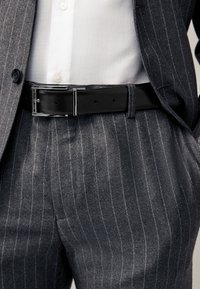 Szary garnitur w cienkie prążki z teksturowanym czarnym skórzanym paskiem. Garnitur ma subtelny połysk, pod marynarką widoczna biała koszula.