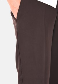 Pantaloni in tessuto marrone scuro con una texture liscia, caratterizzati da una vestibilità slim e dettagli sottili lungo la cucitura laterale.