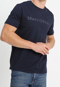 Tummansininen puuvillapaita, jossa on teksti "Marc O'Polo" kontrastivärisessä harmaassa. Klassinen pyöreä kaula-aukko ja lyhyet hihat.